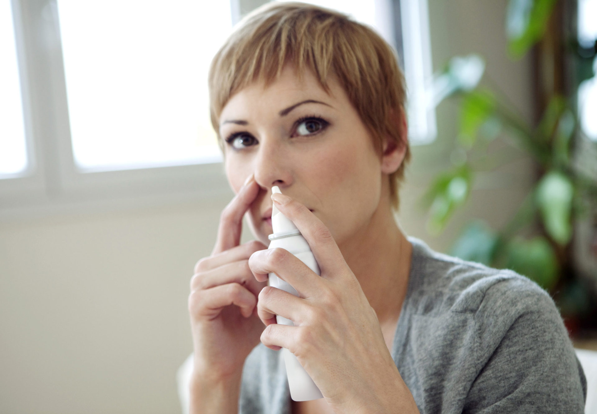 A woman with short blonde hair, wearing a gray sweater, uses a nasal spray with Aptar Pharma’s Bag-On-Valve technology, pressing the spray to her nose in a well-lit room with plants in the background.