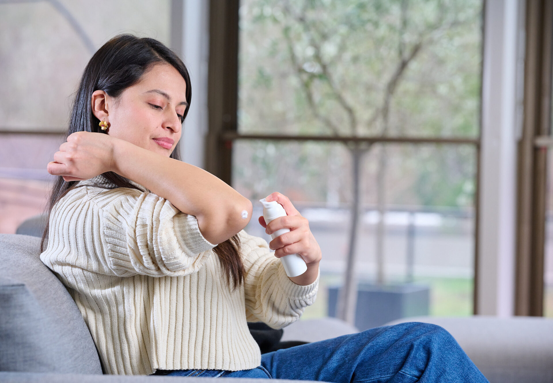 Woman with long brown hair seated in chair dispenses dermal cream on elbow from Aptar Airless+ drug delivery system