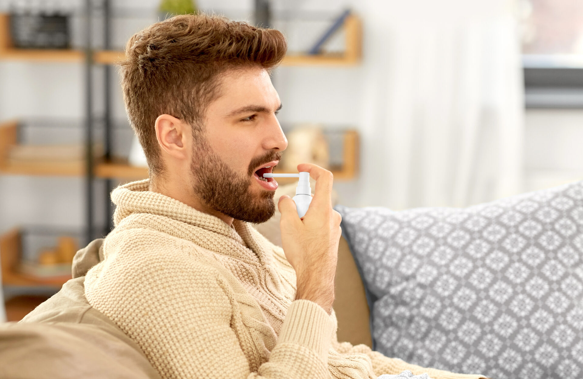 Bearded man in sweater sitting on sofa at home holds Aptar Pharma oral drop dispenser to his mouth delivering VMS product.